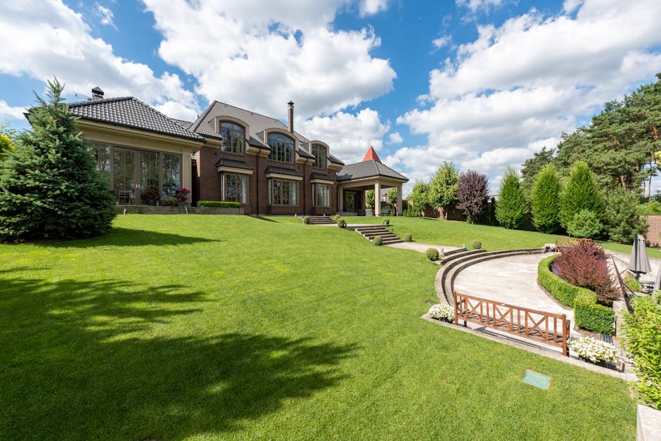 A spacious, well-maintained garden featuring a lush, green lawn with neatly trimmed grass and a curved stone pathway leading up to a modern, two-storey house with large windows, a tiled roof, and a sm